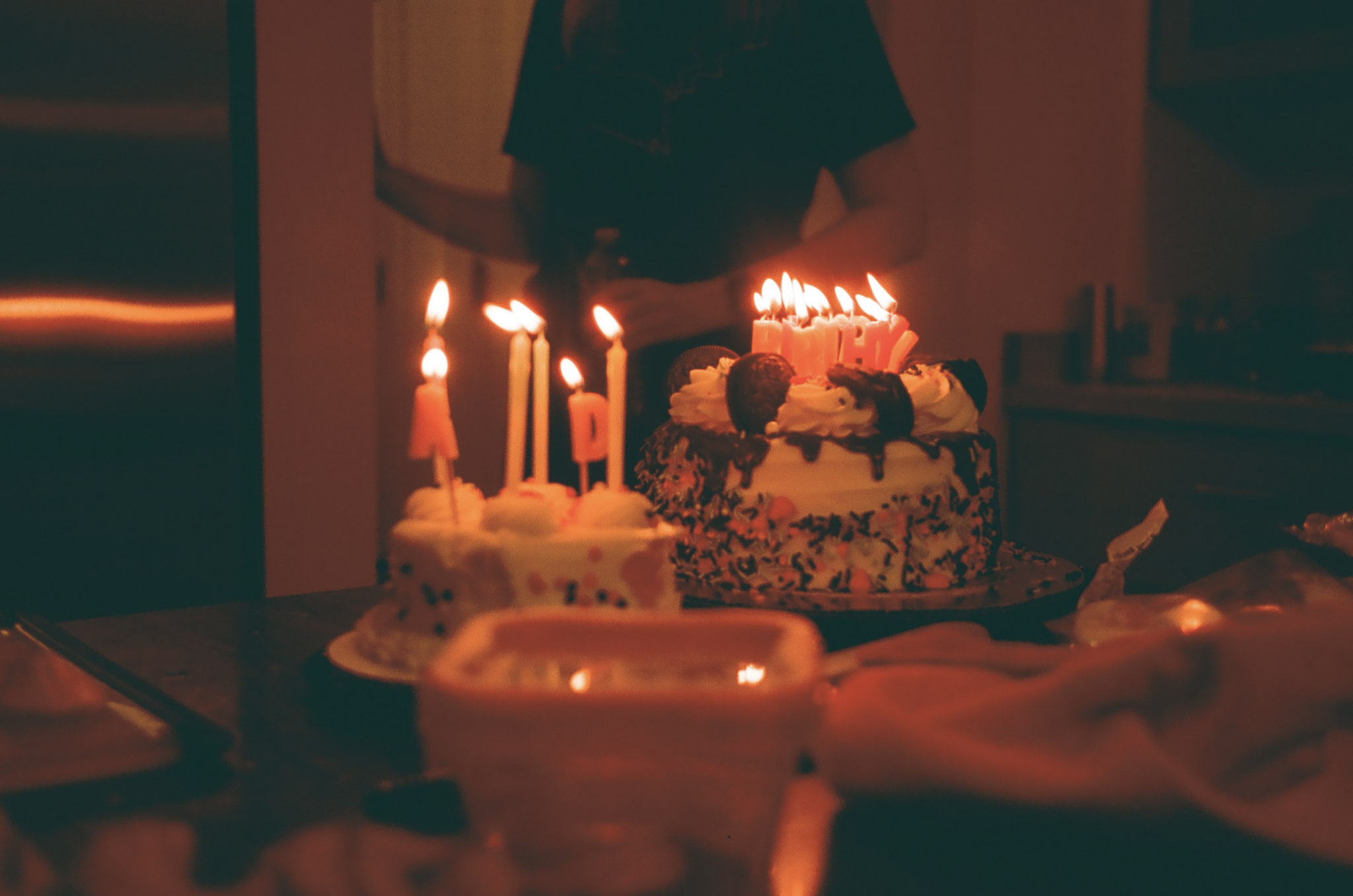 2 birthday cakes on a counter with many blazing candles. They spell birthday.