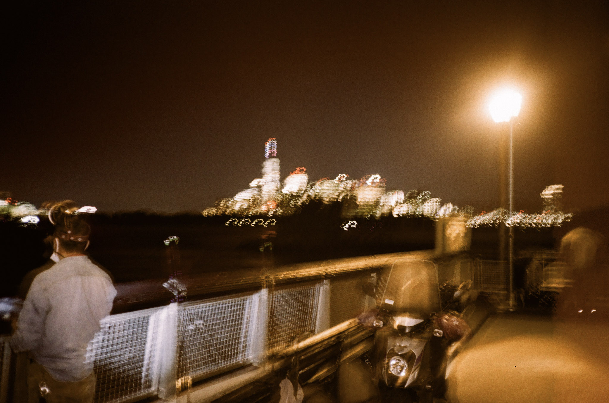 A blurry late night photo off the docks in Redhook looking up to Manhattan. There are a few fishing poles set up and a guy is reeling on in.