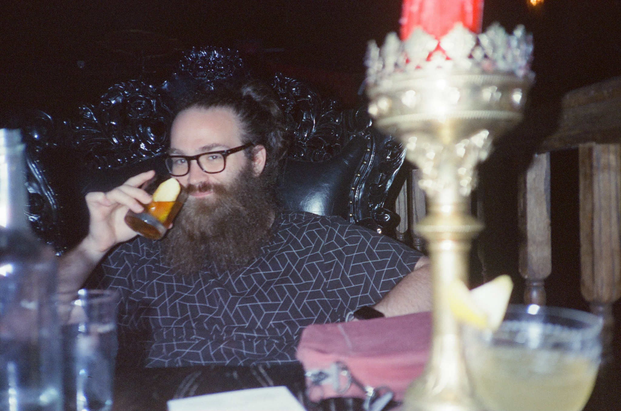 Jake holding a cocktail sitting in a large arm chair with a big candle in the foreground