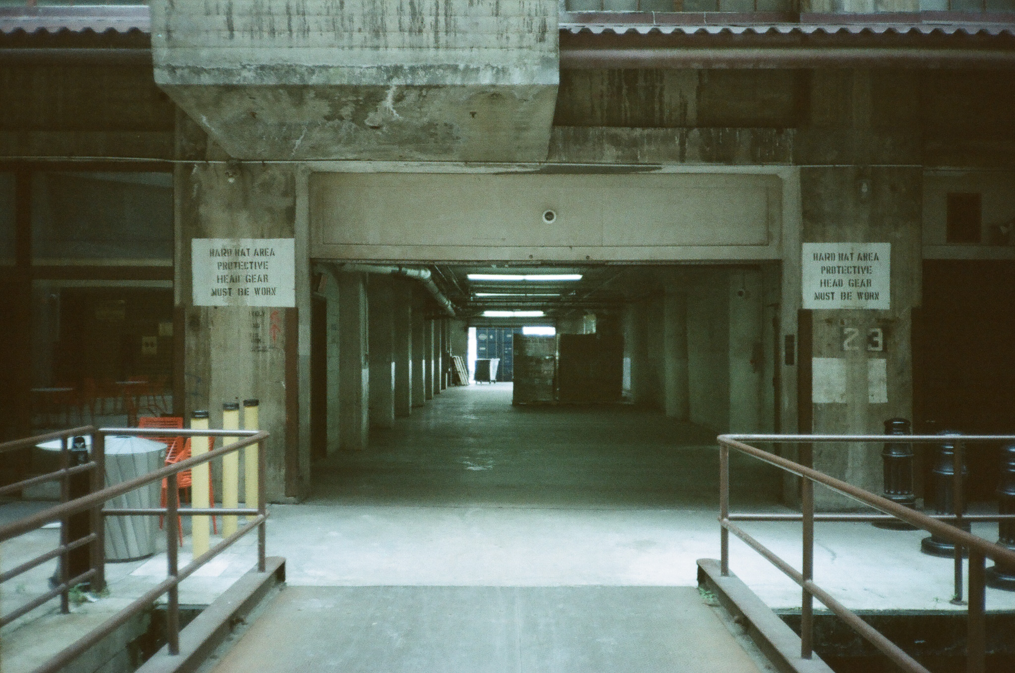 A long concrete hallway with stuff on pallets. Spray painted signage indicates hard hats are required.