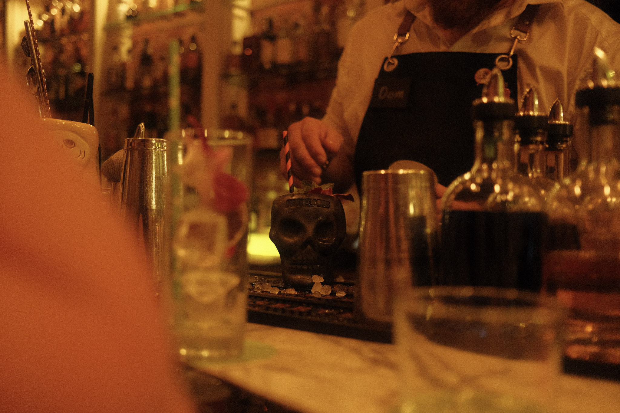 A low shot of a bar at night. The bar is cluttered with glasses, shaking tins, and bitters. In the center the bar tender is finishing a tiki cocktail in a skull glass.