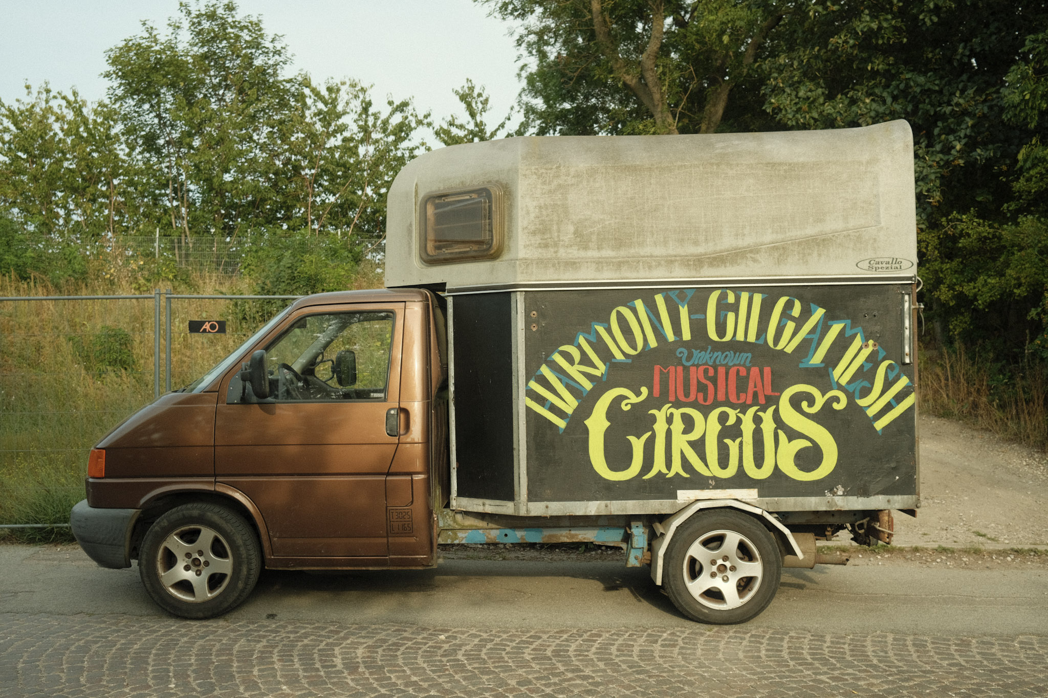 An old brown truck with the bed replaced with a covered trailer. The words 'HARMONY-GILGAMESH Unknown MUSICAL CIRCUS' are painted on in yellow and blue.