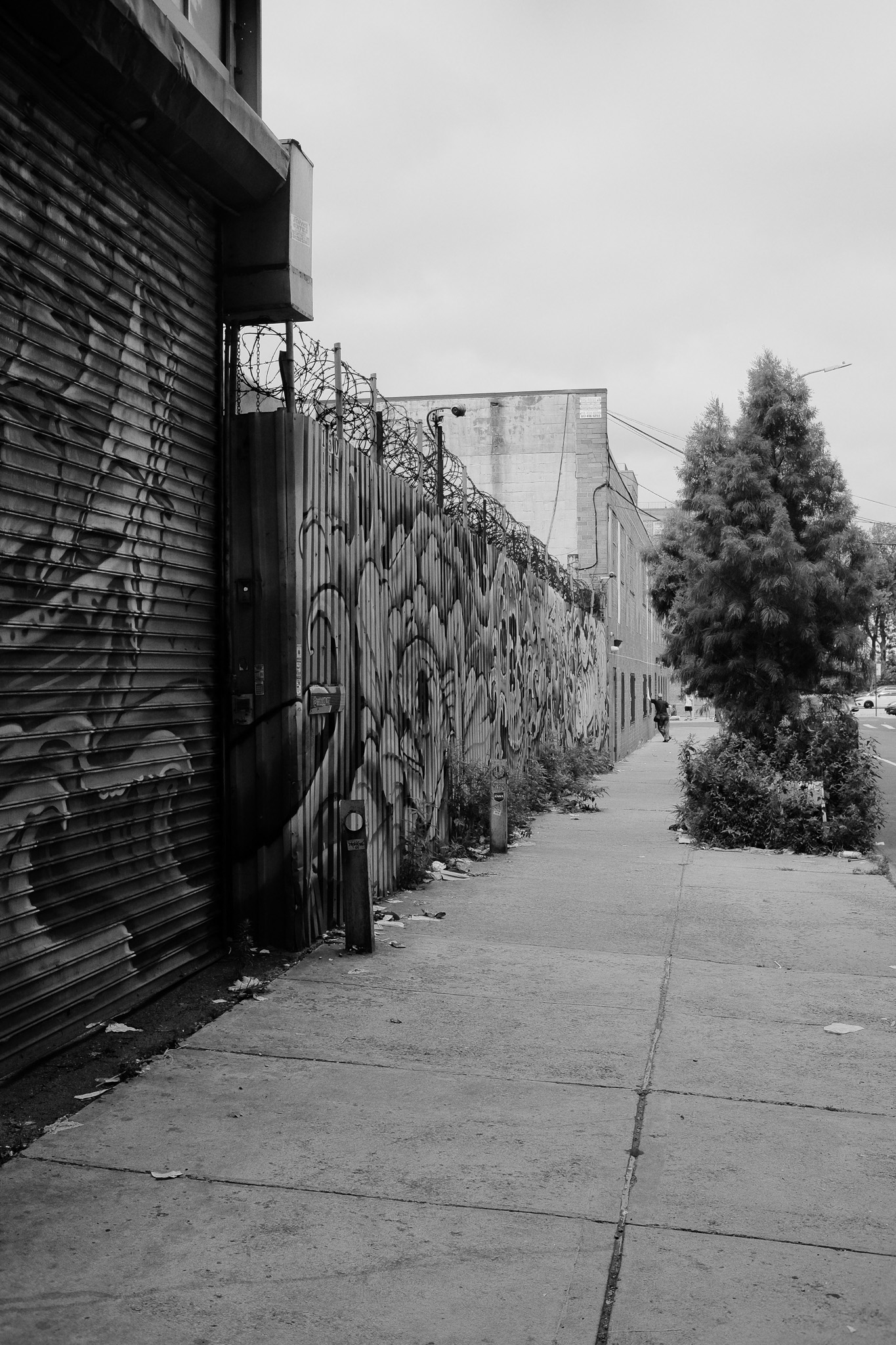 A bunch of graffiti on a bent metal fence topped by razor wire on a street in Brooklyn