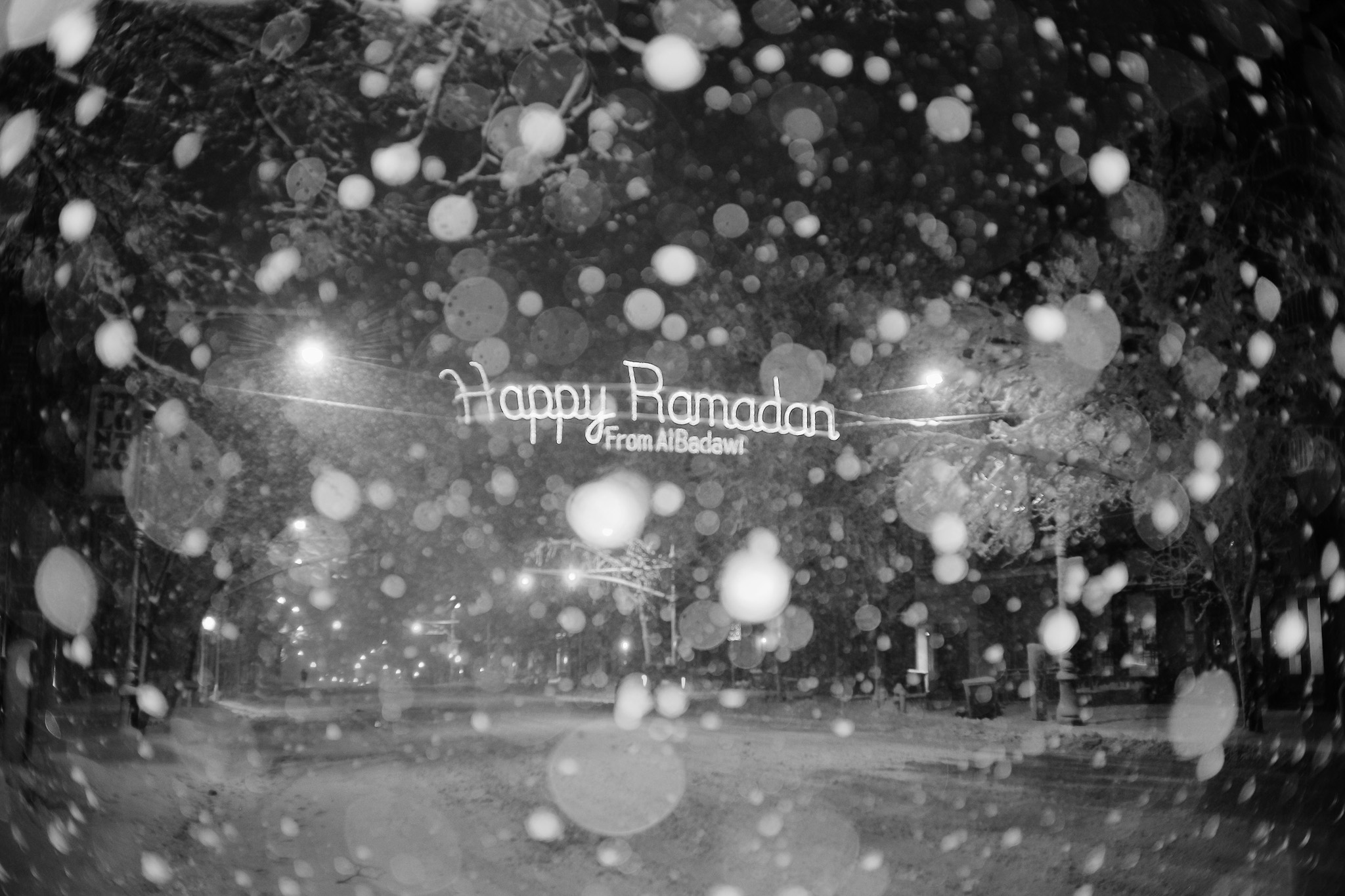 A lit up sign hanging over a wide street that says 'Happy Ramadan from AlBadawi' as shot through a snowy night.
