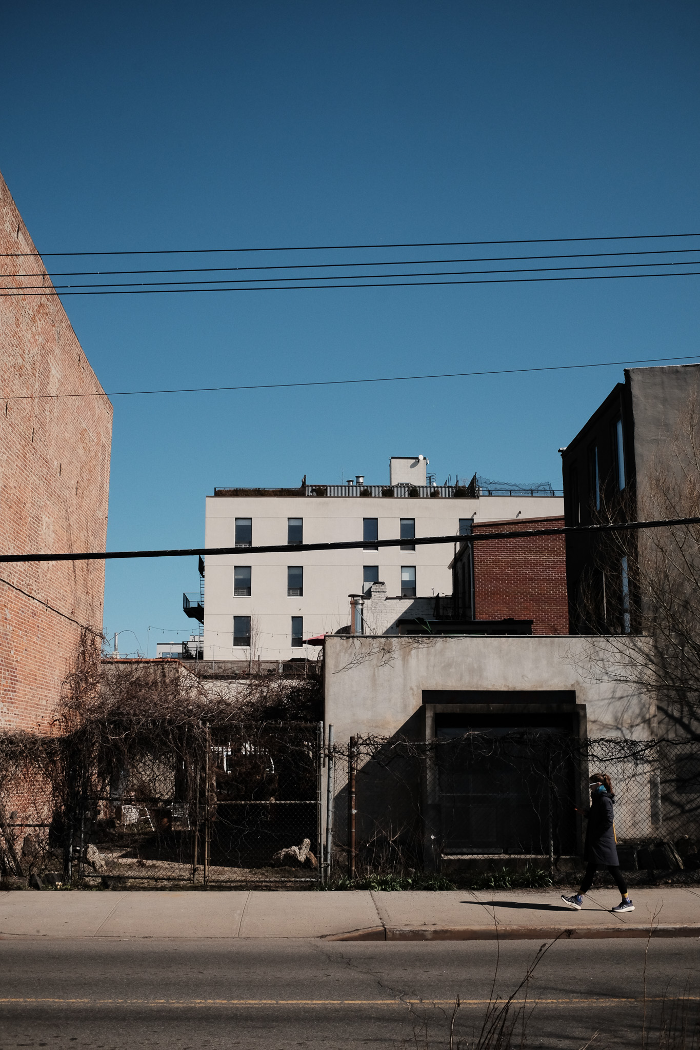 Several old brick buildings with a chain fence in front of them. A woman walks on the sidewalk in front..