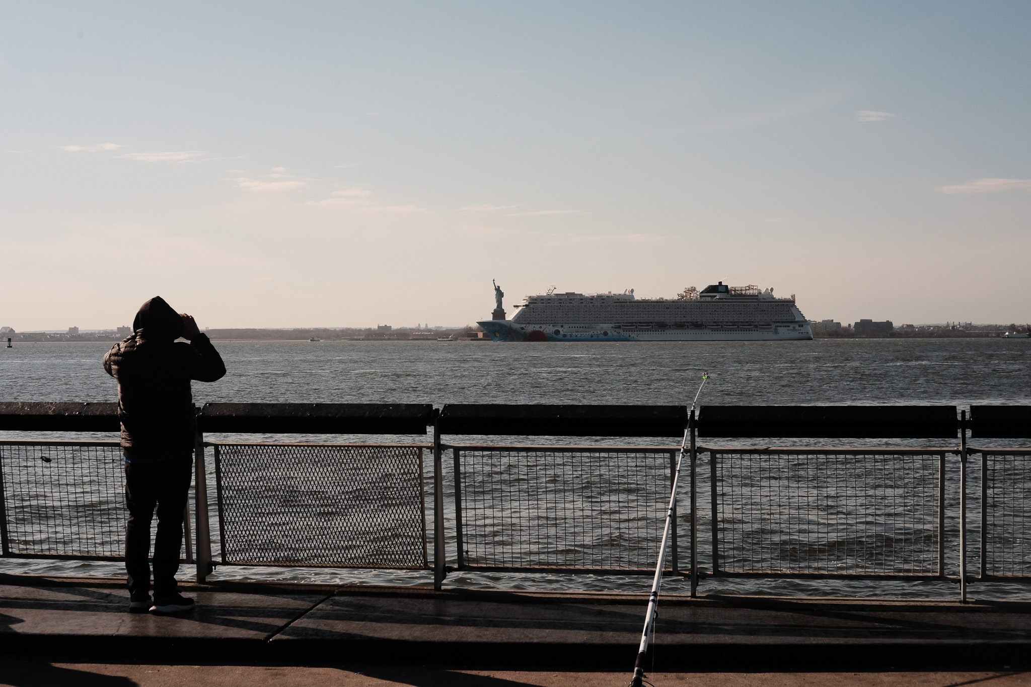 In the distance, a cruise ship crosses in front of the Statue of Liberty. A man watches next to a fishing rod from the pier.